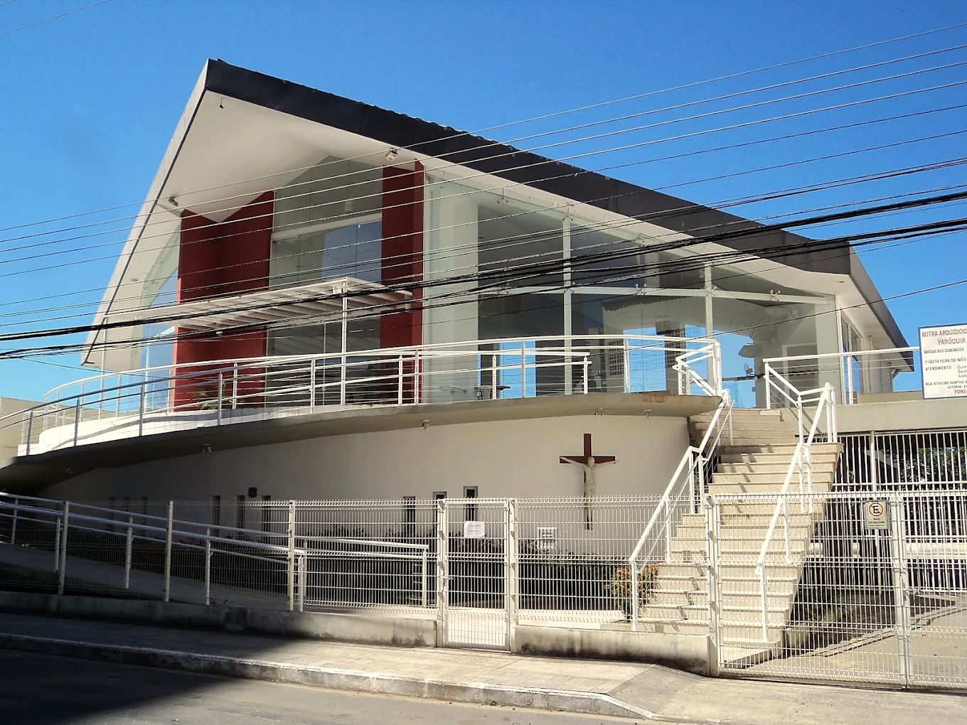 Fachada da Paróquia Santa Luzia: prédio branco com colunas vermelhas, escadaria e crucifixo, sob o céu azul.