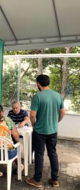Atendimento sob a tenda: pessoas sentadas às mesas e um voluntário de pé, vistos de longe.